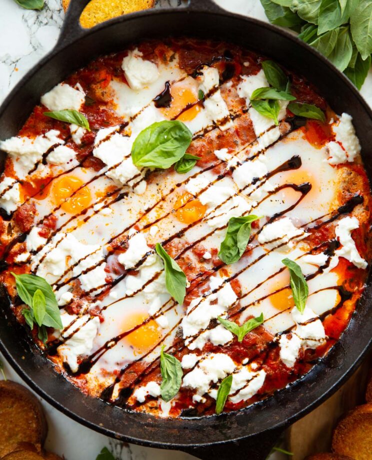 overhead shot of caprese baked eggs in skillet surrounded by toast and fresh basil
